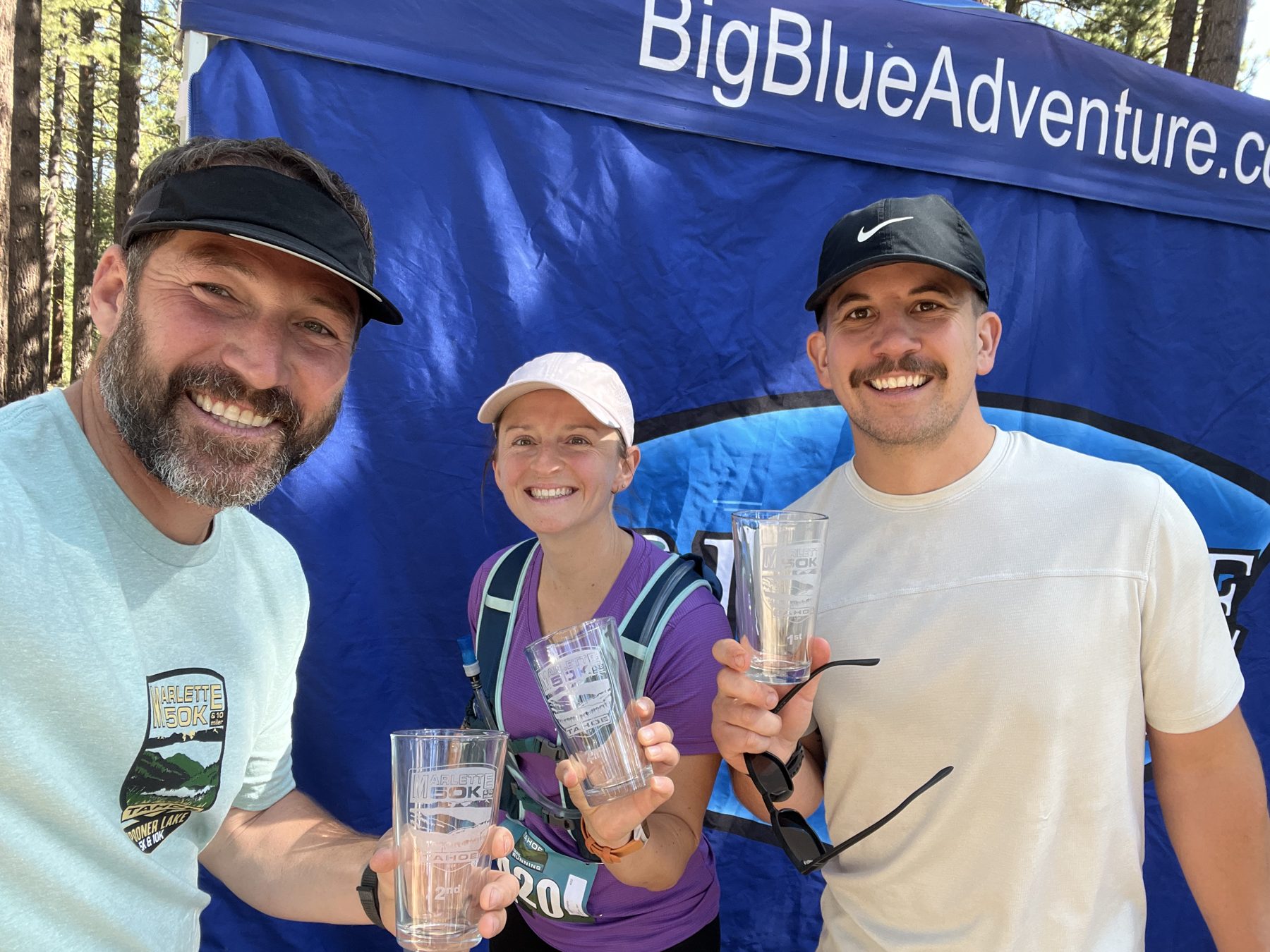 Three finishers holding etched pint glass awards in front of the Big Blue Adventure banner